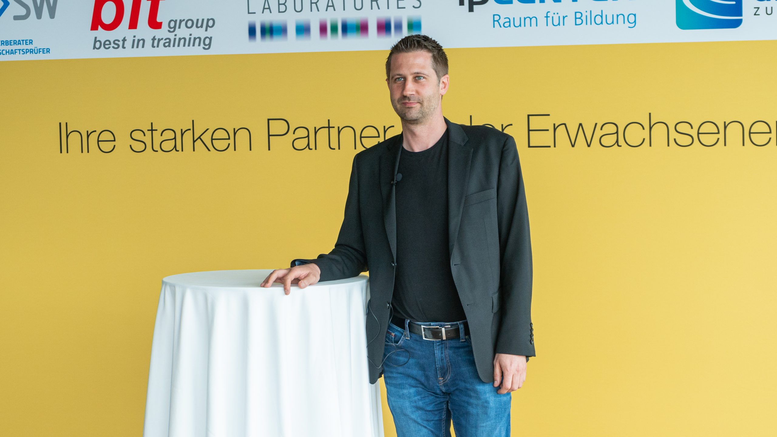 Christoph Wirl steht an einem Tisch vor der Plattform Erwachsenenbildung Messewand